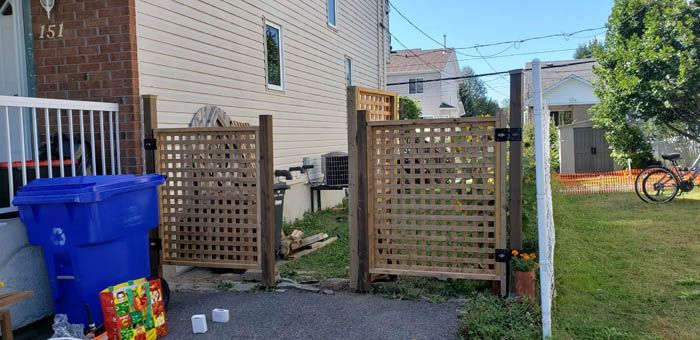 Deux portails en treillis de bois installés entre des poteaux dans une allée résidentielle, avec une poubelle bleue au premier plan.