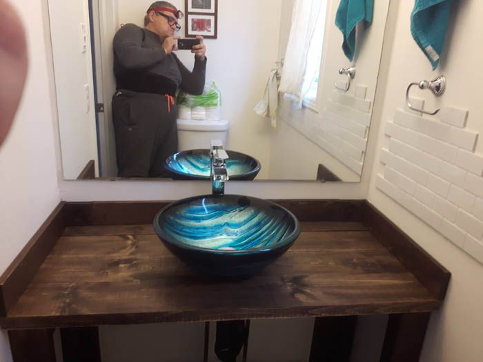 Une personne prend un selfie devant un miroir dans une salle de bains avec un meuble-lavabo en bois rustique et un lavabo en verre bleu vif à motifs tourbillonnants.
