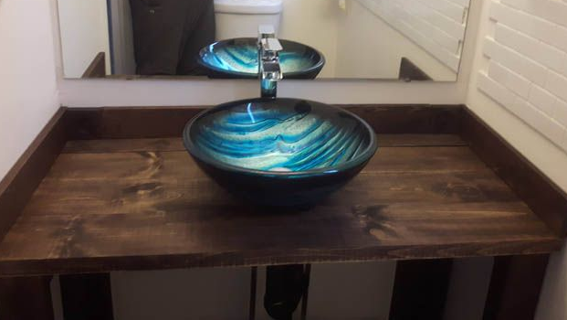 Dans une salle de bains avec miroir mural, un lavabo vasque en verre bleu et turquoise est posé sur un meuble-lavabo rustique en bois foncé.