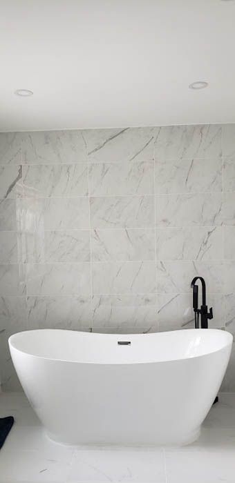 Une baignoire îlot blanche moderne, adossée à un mur de marbre blanc, avec un robinet noir fixé au sol.