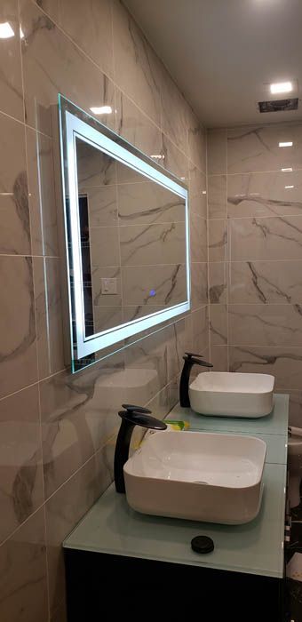 Un meuble-lavabo moderne avec deux vasques carrées blanches, des robinets noirs et un miroir mural rectangulaire éclairé.
