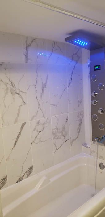 Une cabine de douche moderne avec un mur à motif marbre blanc, une baignoire blanche et un panneau de douche argenté illuminé d'une lumière bleue.