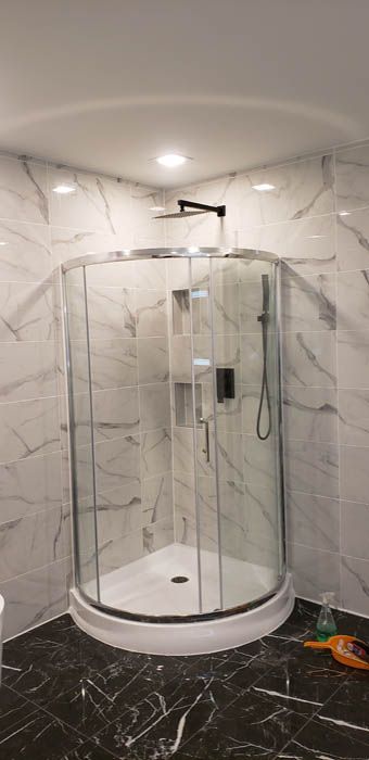 Une cabine de douche d'angle avec portes vitrées incurvées, murs à motifs de marbre blanc et sol à motifs de marbre noir.