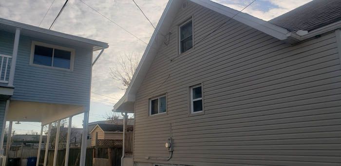 Une maison bleu clair, à gauche, se dresse à côté d'une maison beige à deux étages au bardage horizontal, sous un ciel nuageux.