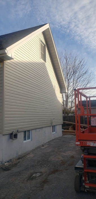 Un revêtement extérieur en vinyle clair recouvre le côté d'une maison avec deux petites fenêtres au sous-sol, à côté d'une nacelle élévatrice orange.