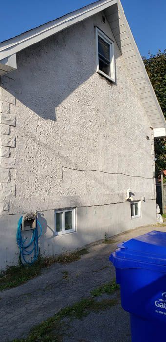 Vue latérale de l'extérieur d'une maison avec un enduit clair, une fissure horizontale visible, deux fenêtres au sous-sol et un tuyau d'arrosage.