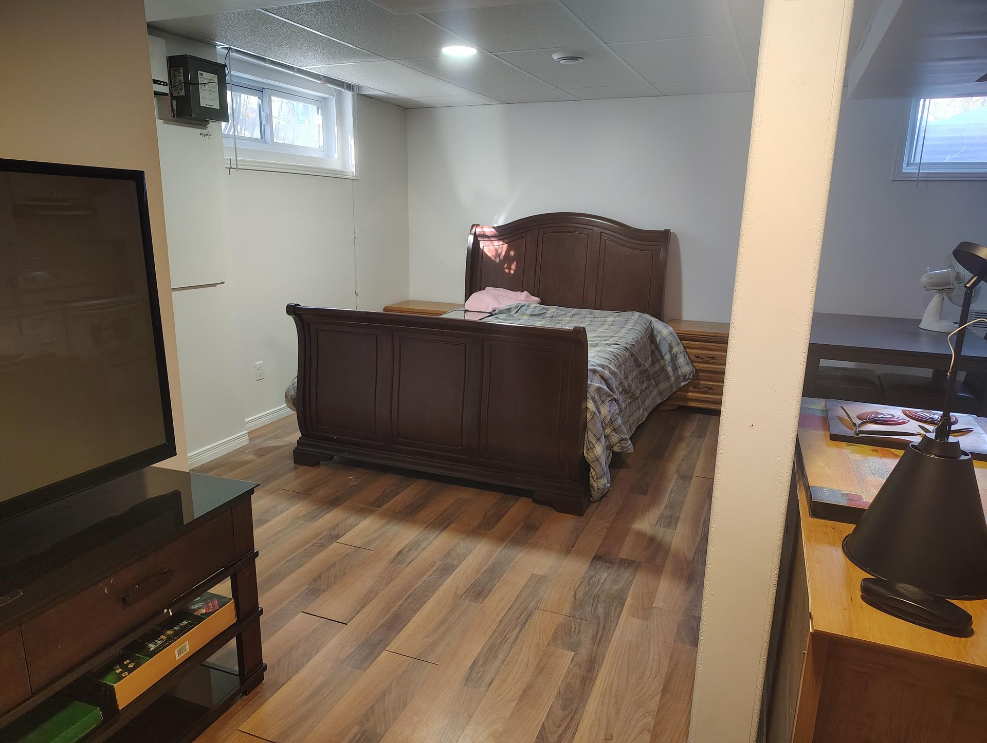Une chambre au sous-sol avec un lit traîneau en bois foncé, un sol imitation bois, deux petites fenêtres et un meuble TV.