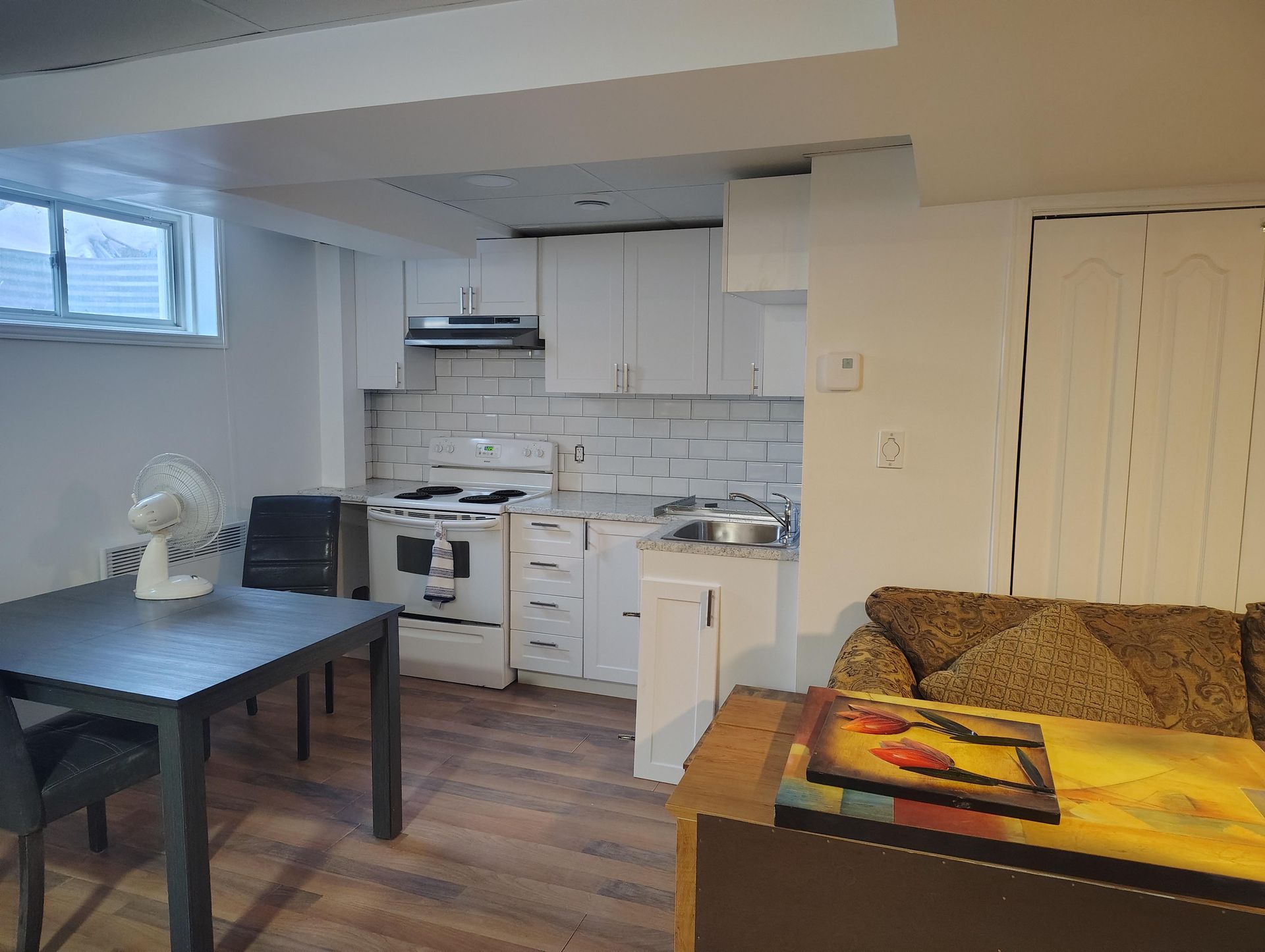 Un petit appartement en sous-sol comprenant une kitchenette avec des placards blancs, une petite table à manger et un canapé beige.