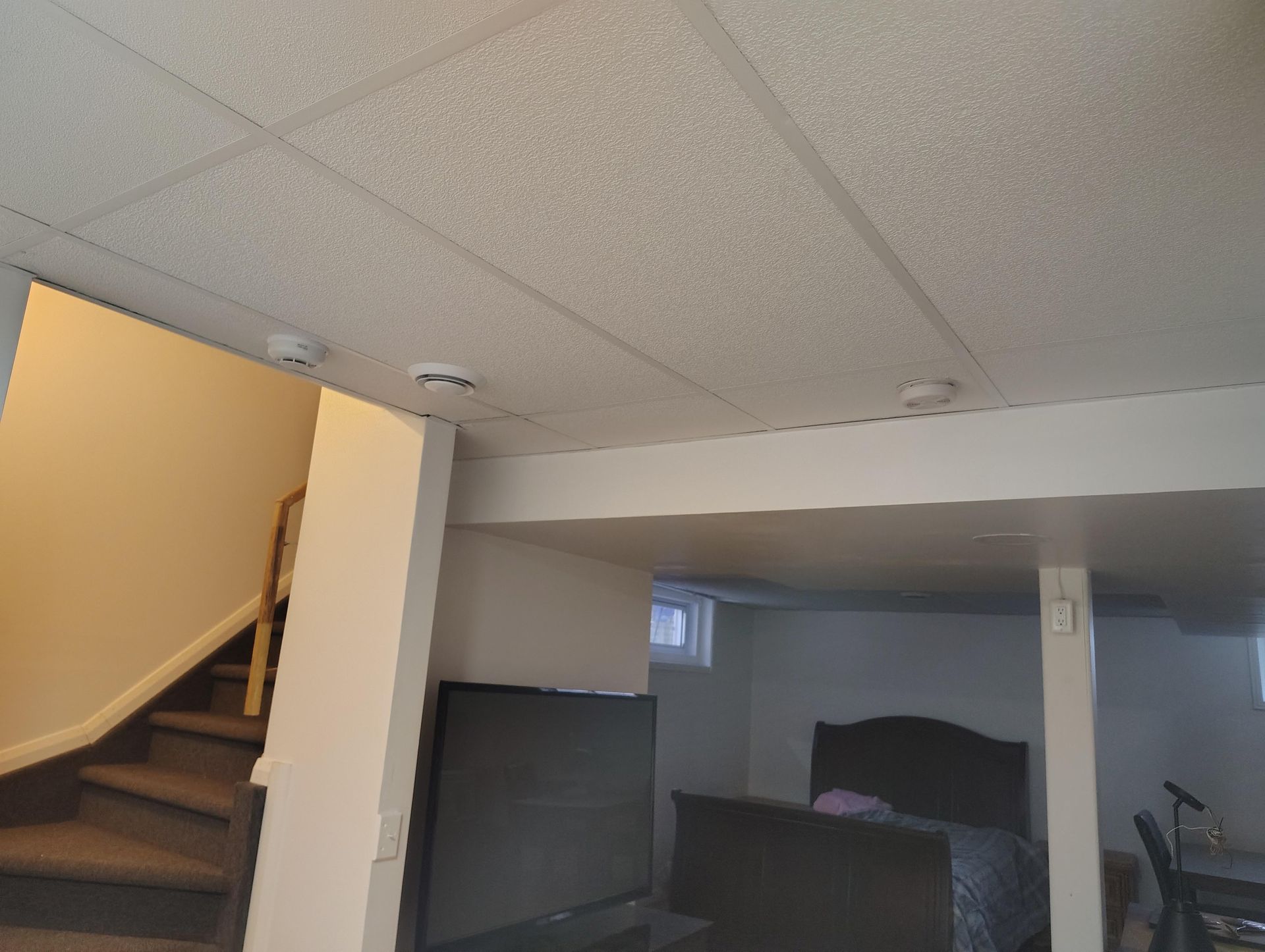 Un sous-sol d'habitation montrant un escalier à gauche, une télévision au premier plan et une chambre à coucher à l'arrière-plan.