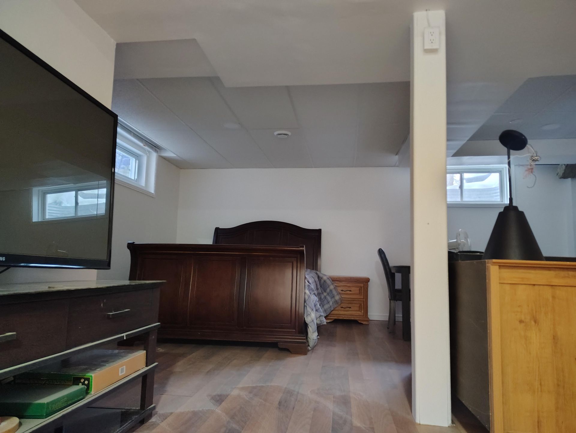 Une chambre au sous-sol avec un lit en bois, une table de chevet et un bureau, vue depuis une pièce avec une télévision et un pilier de soutien.