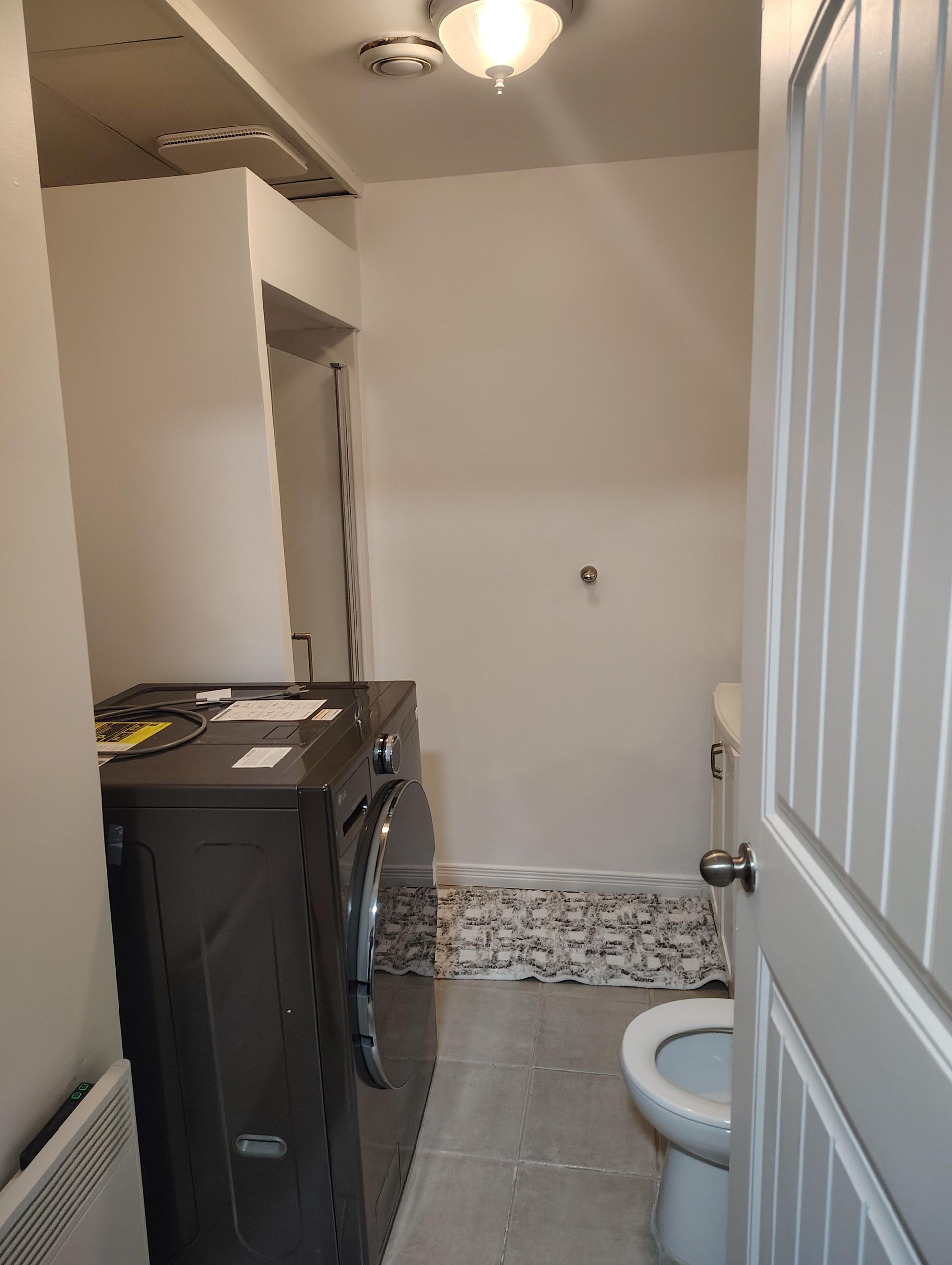 Une petite salle de bains utilitaire comprenant une machine à laver gris foncé, des toilettes blanches et un sol carrelé.