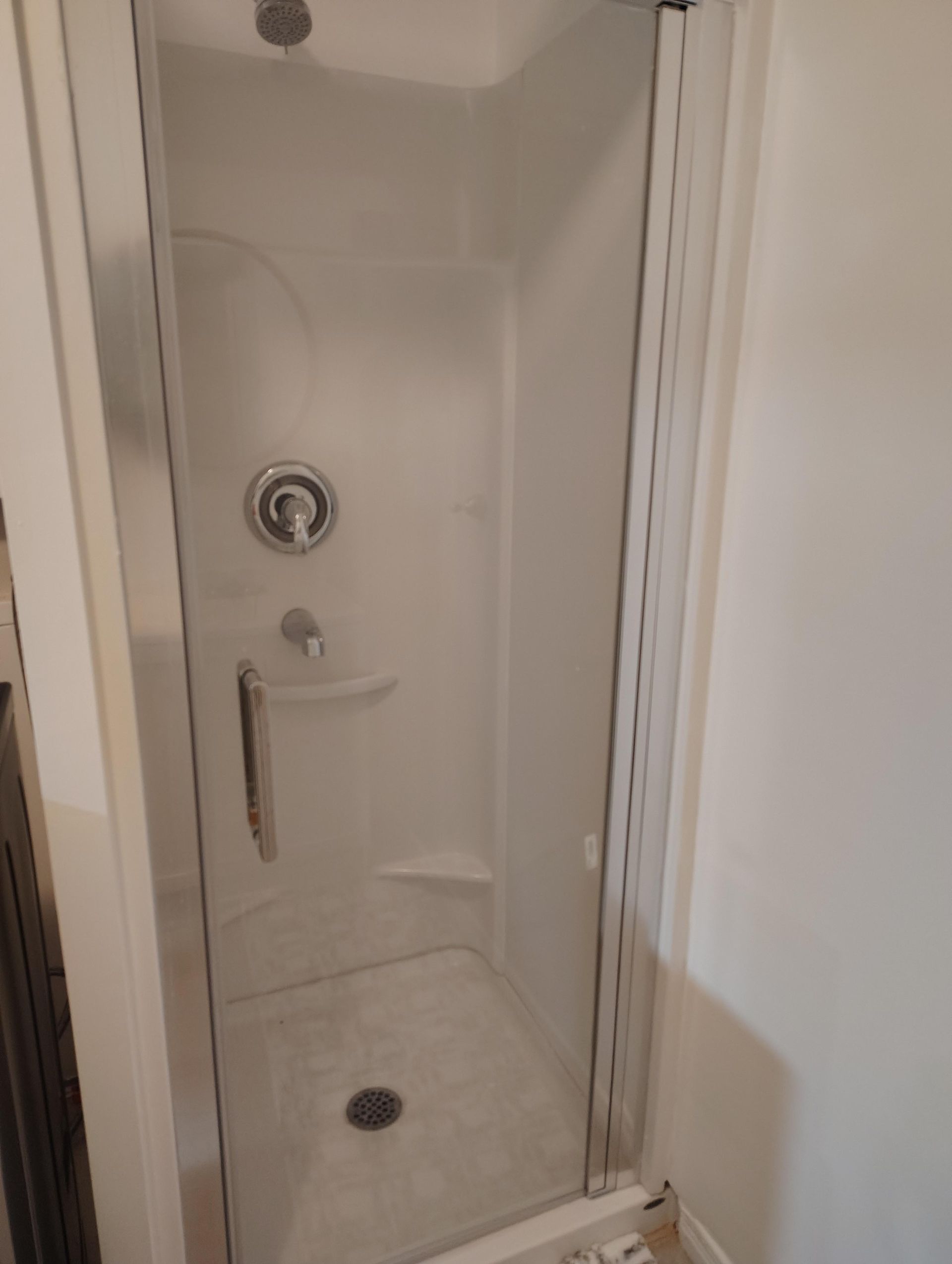 Une petite cabine de douche blanche préfabriquée avec une seule porte en verre et des accessoires argentés dans une pièce simple.
