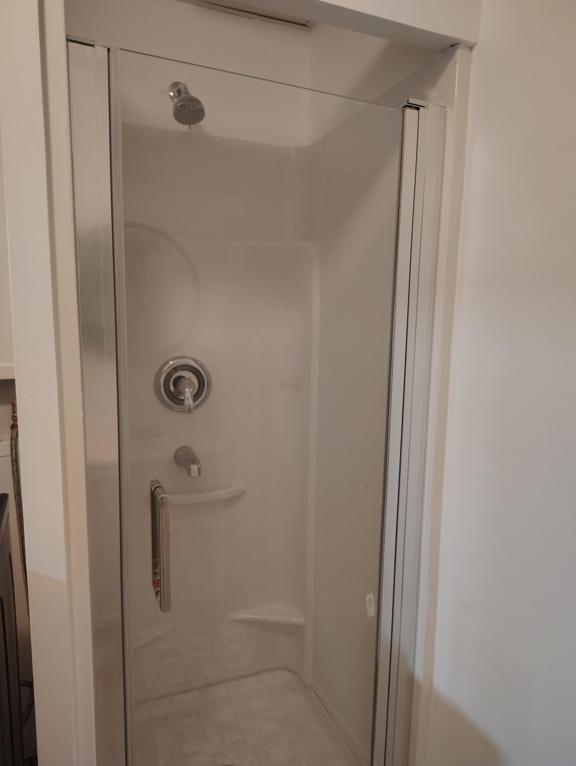 Une douche blanche de type cabine avec porte en verre, pommeau de douche, robinet et poignée chromés, adossée à un mur uni.