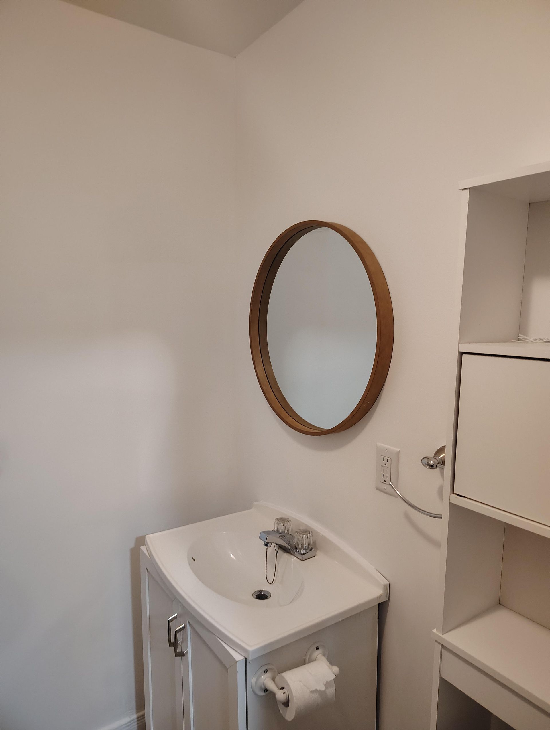 Un meuble-lavabo avec un lavabo blanc, un robinet chromé, un miroir rond à cadre en bois au-dessus et des étagères blanches adjacentes.