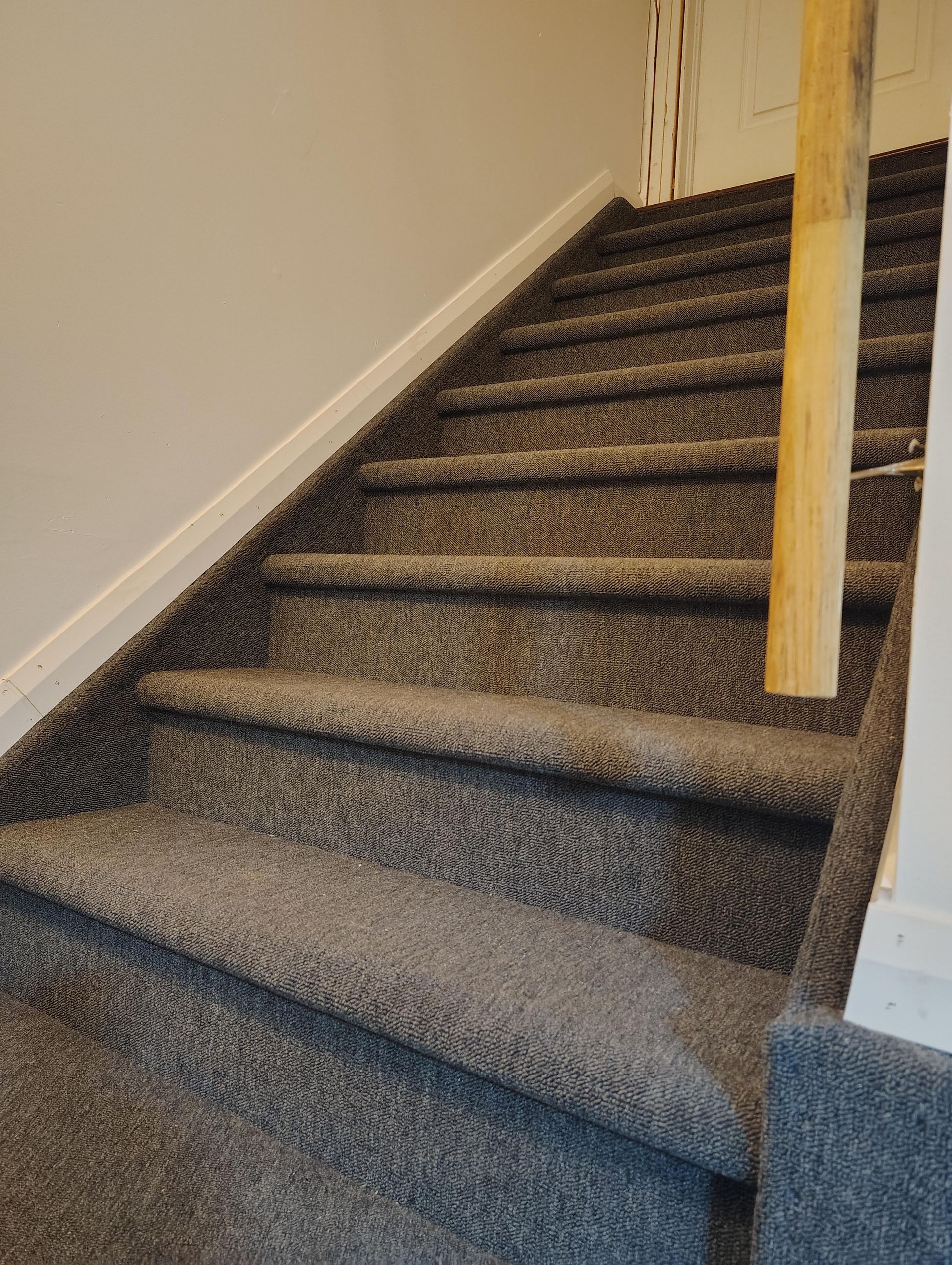 Escalier recouvert d'une moquette en tissu texturé gris foncé, menant vers le haut à côté d'un poteau en bois clair.