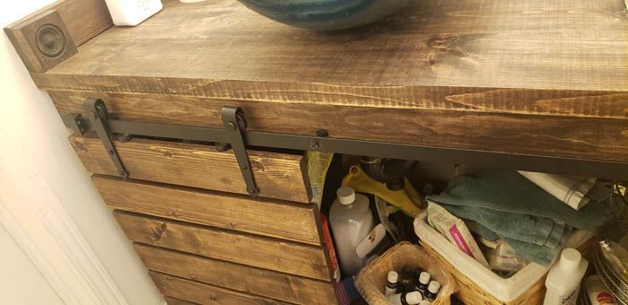 Un meuble-lavabo rustique en bois avec un panneau coulissant de type porte de grange, laissant apparaître une étagère ouverte contenant divers récipients.