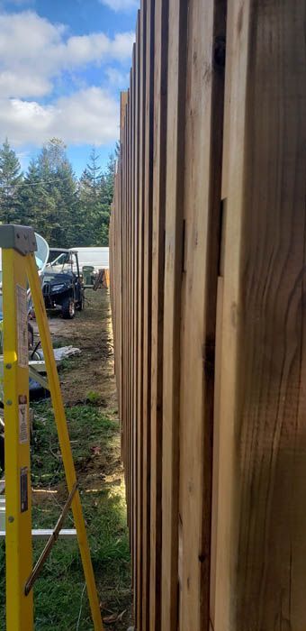 Une échelle jaune se dresse dans une cour en terre battue, à côté d'une haute clôture en bois verticale qui préserve l'intimité, sous un ciel bleu partiellement nuageux.