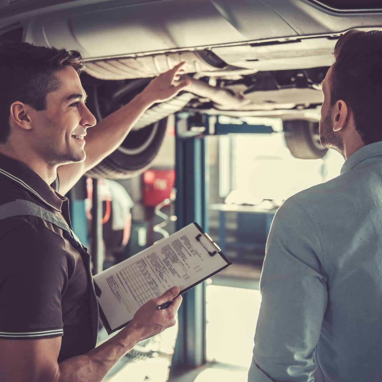 A mechanic in a garage points to the underside of a raised car while explaining a repair to a customer holding a notepad.