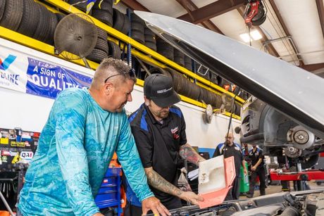 A mechanic gestures to the undercarriage of a car on a lift while explaining repairs to a customer holding a clipboard.