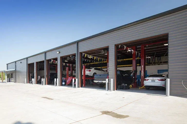 A grey, metal-sided commercial garage with open bays housing several vehicles on lifts under a clear blue sky.