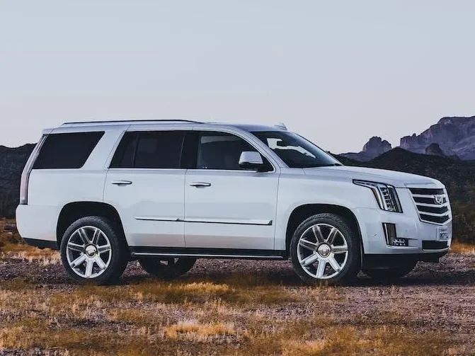 A white Cadillac Escalade SUV parked on a dry, grassy field against a mountainous, dusk-lit horizon.