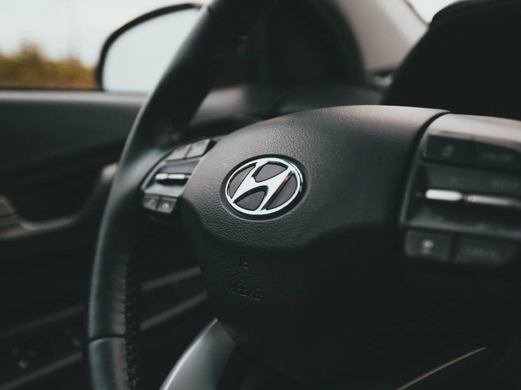 Close-up of a black Hyundai steering wheel with the silver logo in the center, inside a vehicle cabin.