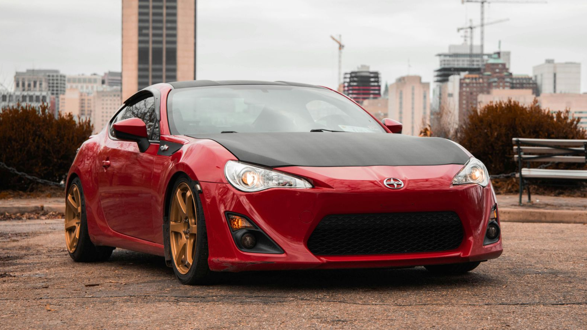 A red sports car with a black hood and gold wheels parked on a paved lot in front of a city skyline.