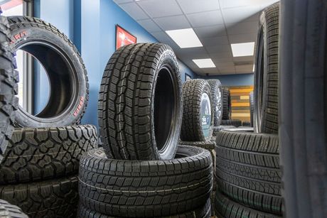 A stack of four black tires with aggressive tread patterns, with one silver five-spoke alloy wheel leaning against them.