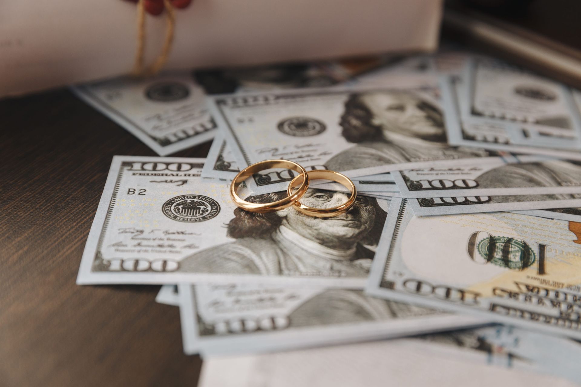 Two gold wedding rings on U.S. dollar bills beside a blurred pen and hand