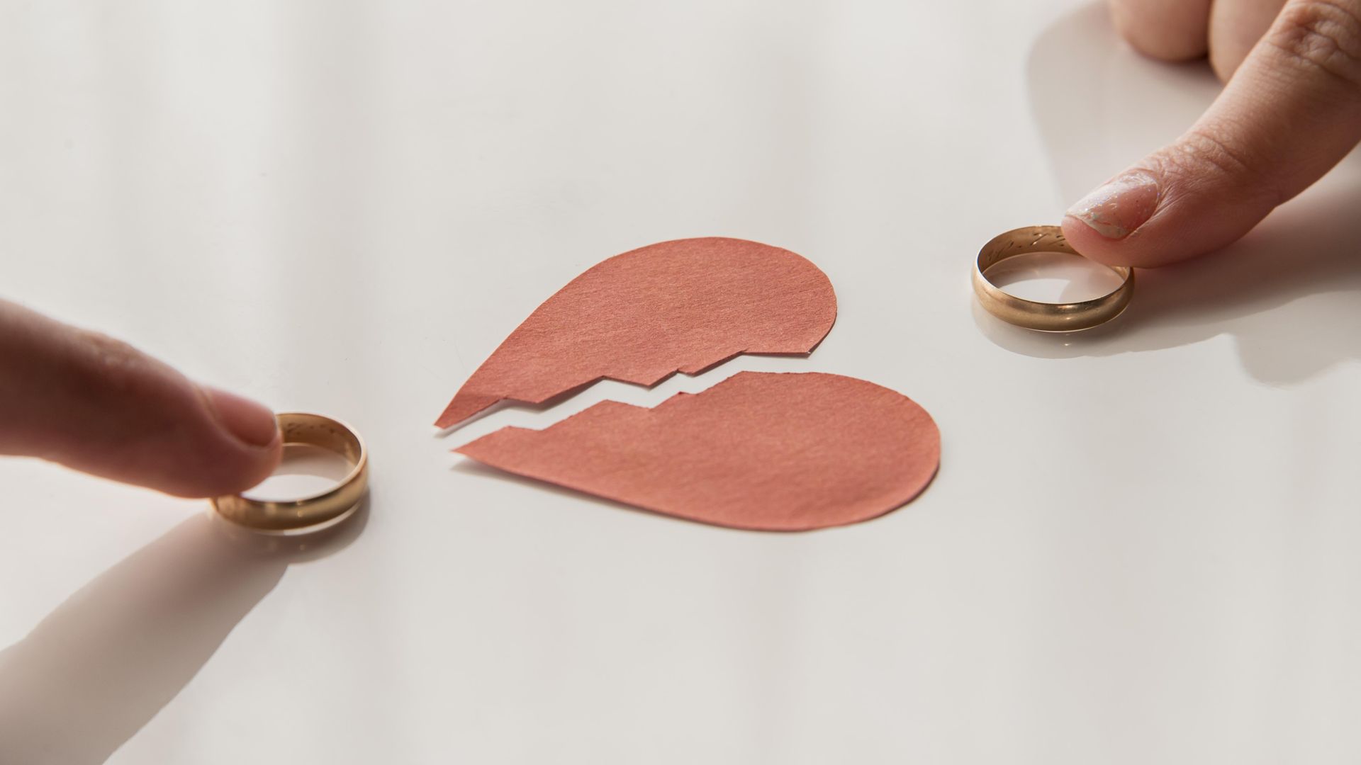 Hands holding two rings around a broken red heart cutout on a white surface