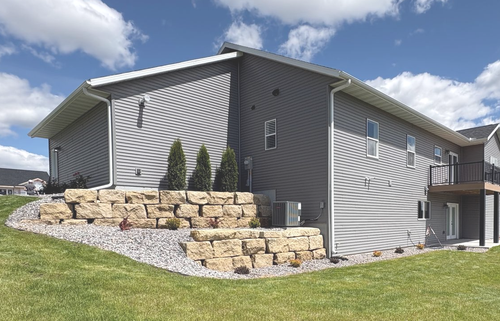 A gray, two-story suburban home with a back deck, stone retaining walls, and a large grassy yard under a blue sky.