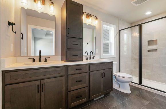 Modern bathroom featuring dark wood cabinetry, a dual-sink vanity, a tiled walk-in shower, and a white toilet.