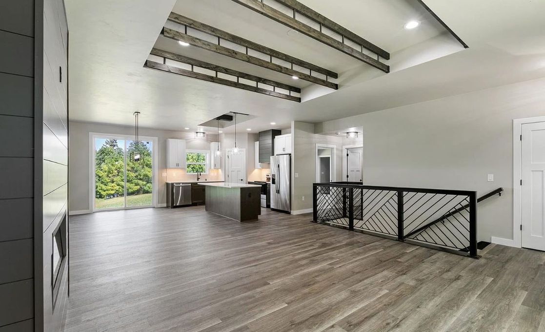 A modern, open-concept kitchen and living area with wood floors, exposed ceiling beams, and a black staircase railing.