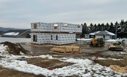 A partially built house with exterior sheathing stands on a snowy, muddy construction site near other homes.