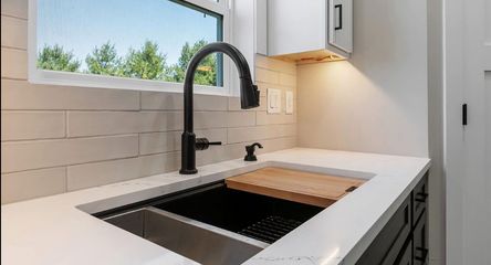 A kitchen sink with a black faucet and a wooden cutting board set into the basin against a tiled backsplash and window.