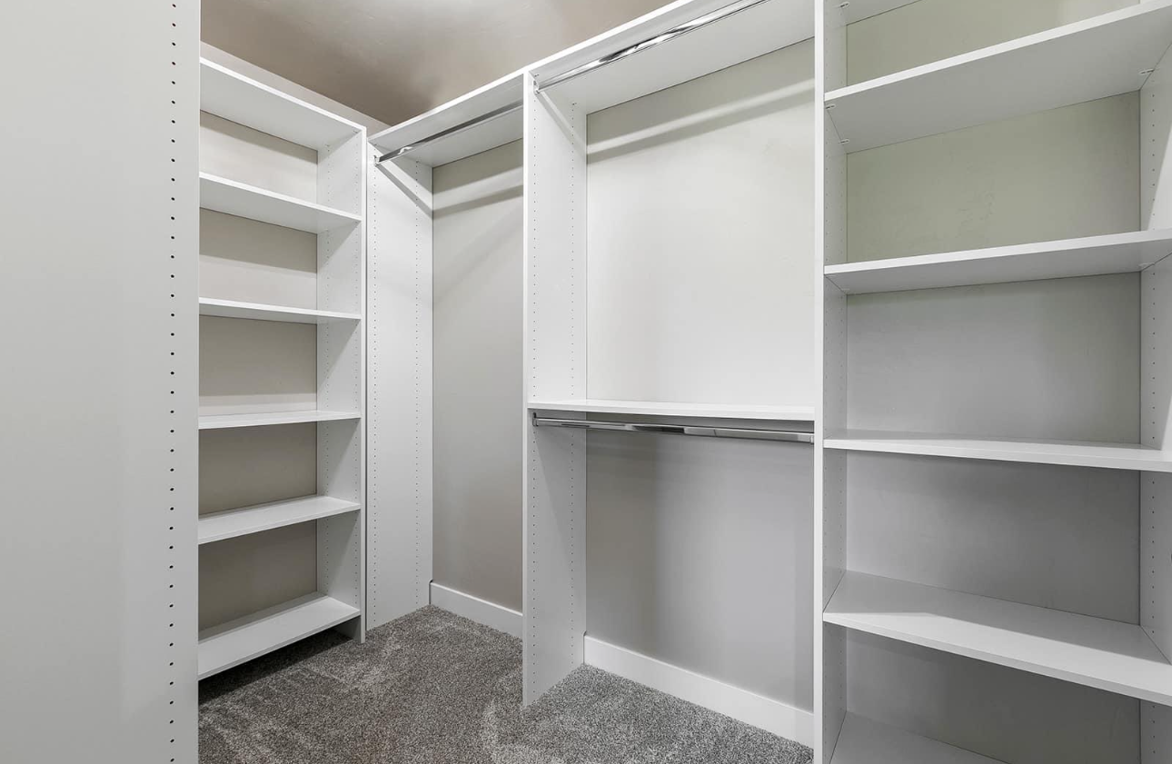 A walk-in closet with white shelving, hanging rods, and grey carpet flooring.