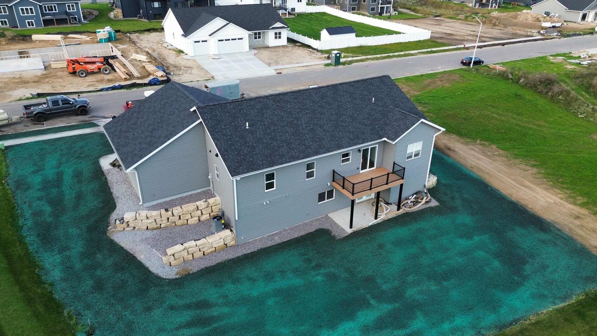 Aerial view of a gray, multi-level suburban home with a dark shingled roof, a back deck, and freshly seeded green lawn.