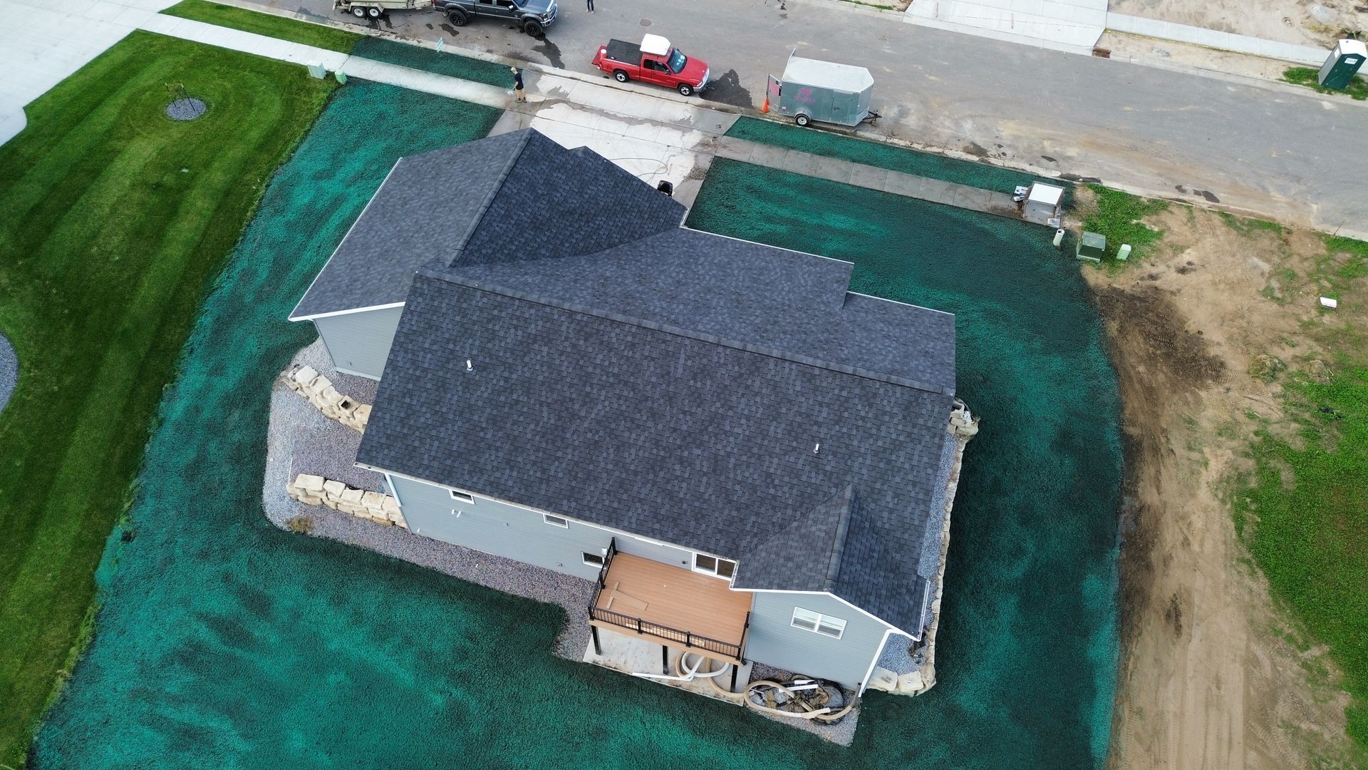 Aerial view of a gray-roofed house surrounded by bright green hydroseeded soil, with a street and construction equipment.