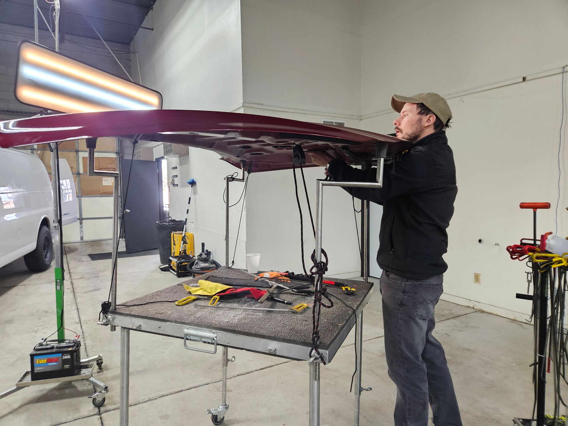 Person in a garage adjusts a maroon car body on a metal frame, with tools and a work light nearby.
