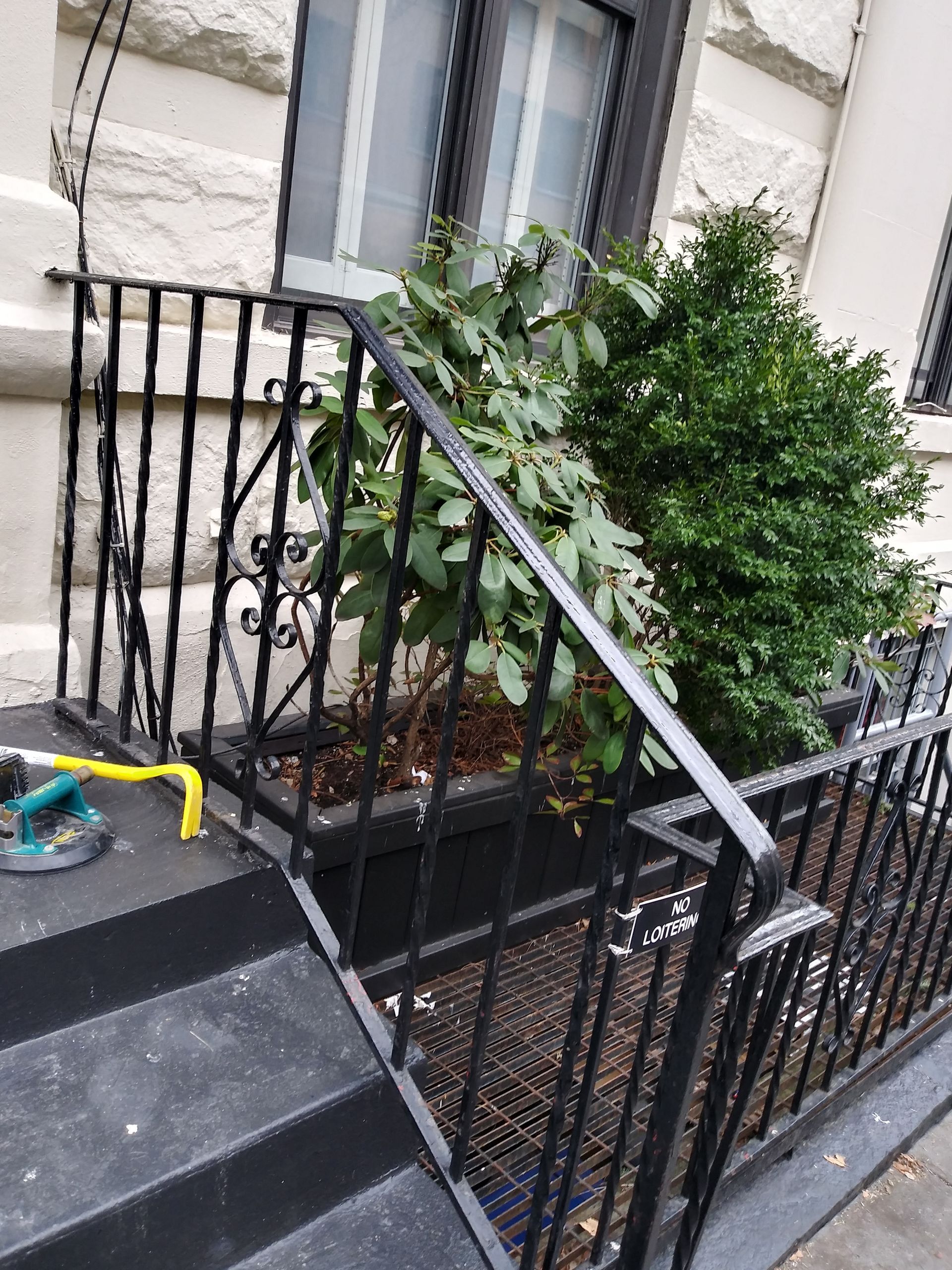 Black metal stair railing beside a stone house, with a green shrub and a yellow-handled tool on the steps.