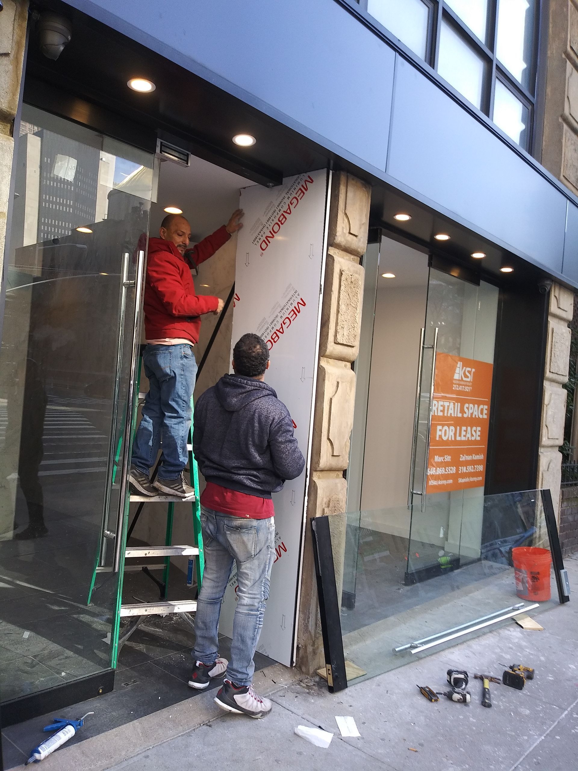 Two workers installing a storefront panel beside a ladder and glass storefront.