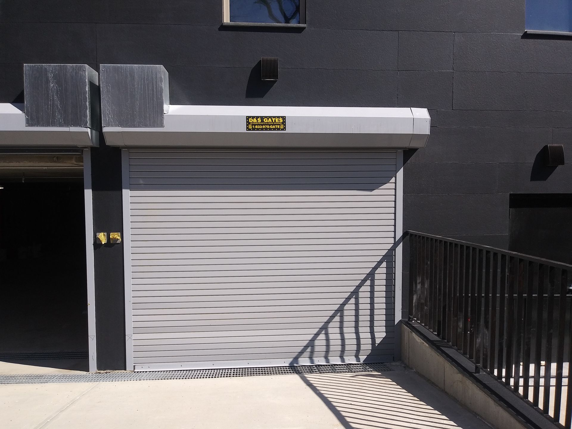 Closed white roll-up garage door on a dark building, with a ramp and railing in front.