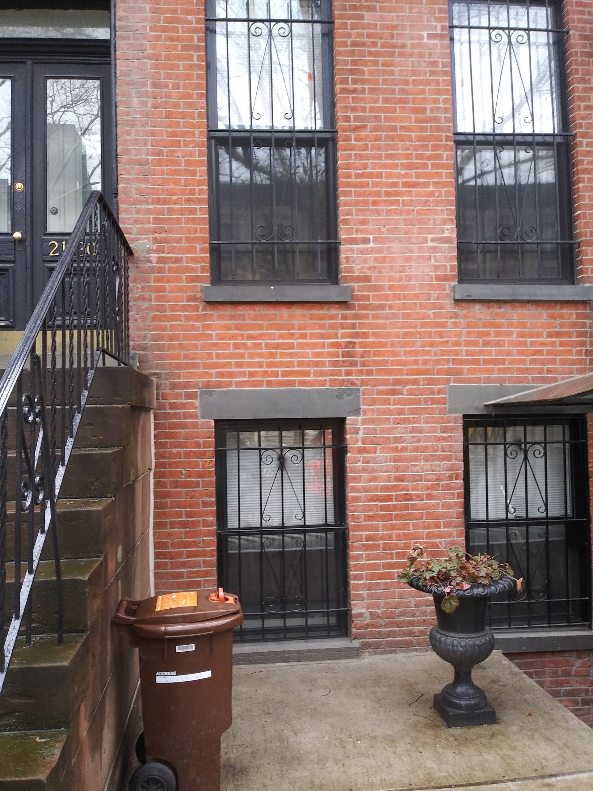Brick townhouse courtyard with black iron stairs, barred windows, a trash bin, and a flower planter