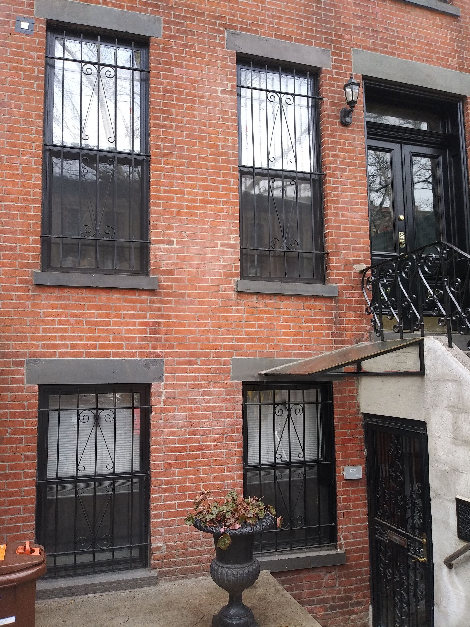 Brick townhouse facade with barred windows, black door, and small potted plant by steps