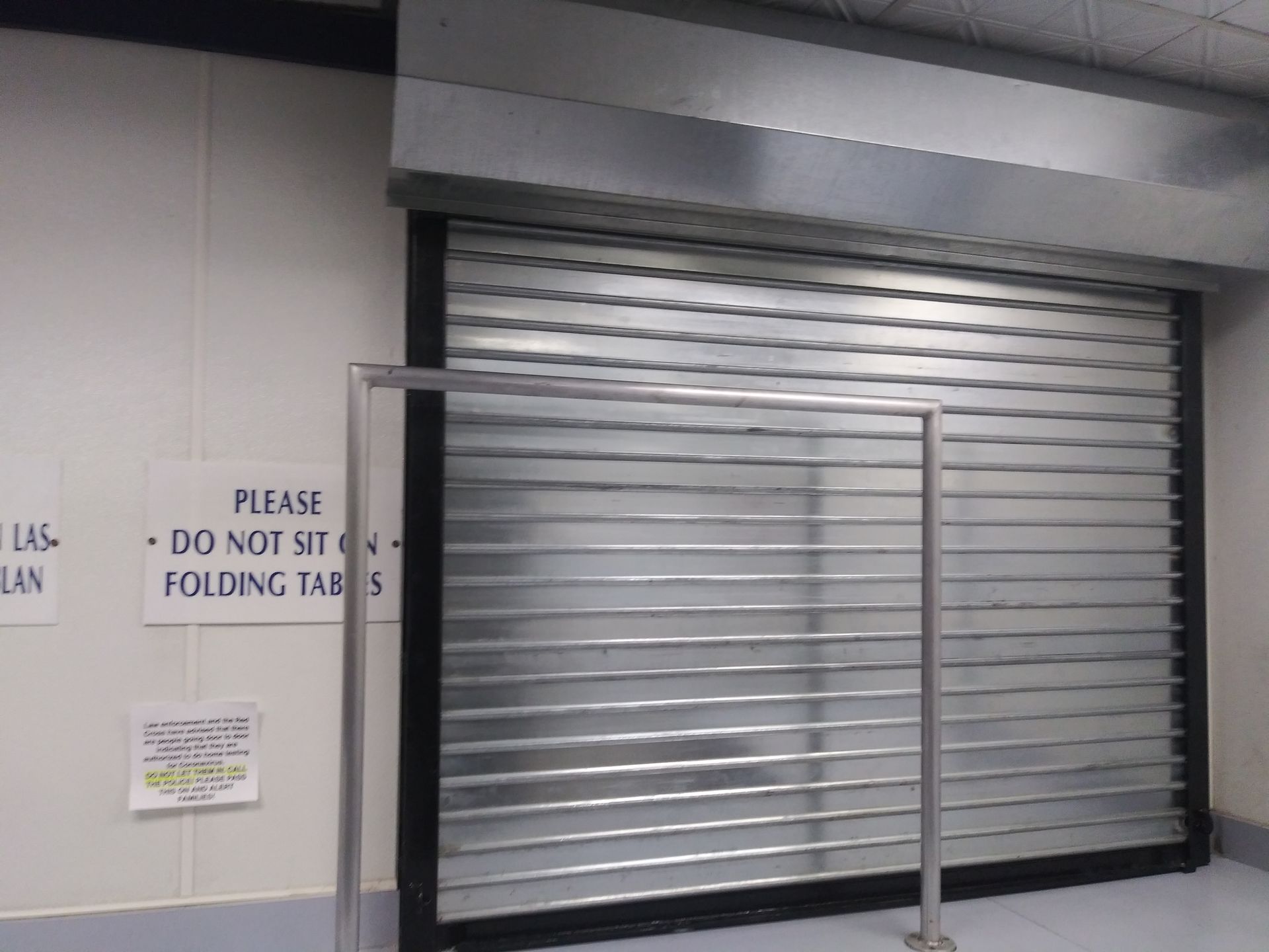 Closed metal security shutter over a storefront window, with a “Do not stack folding tables” sign nearby