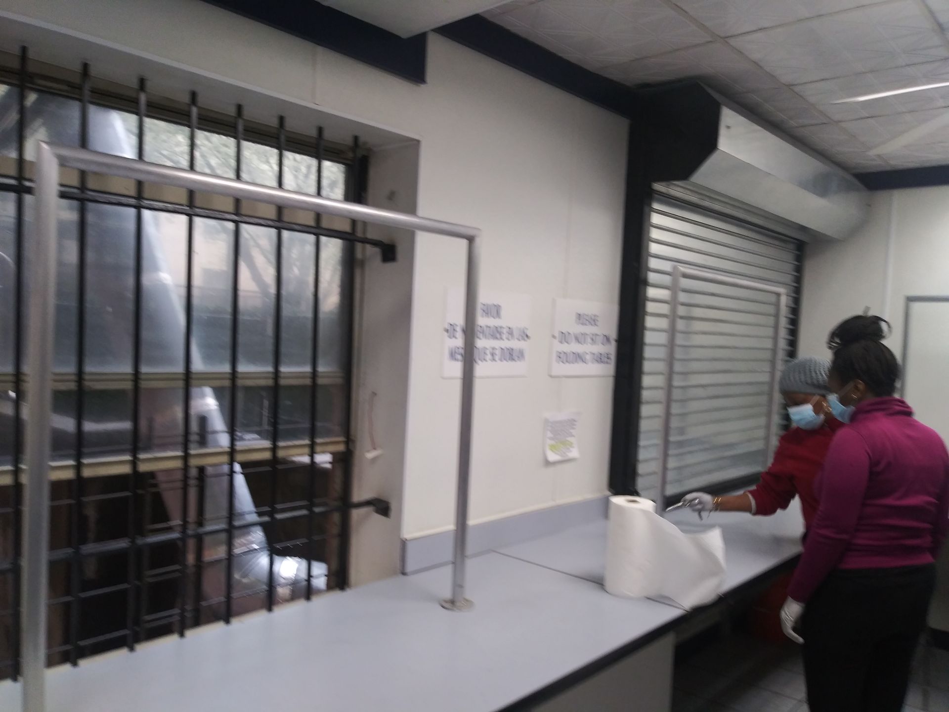 Two masked people stand at a service counter with a pass-through window and bars on the left.
