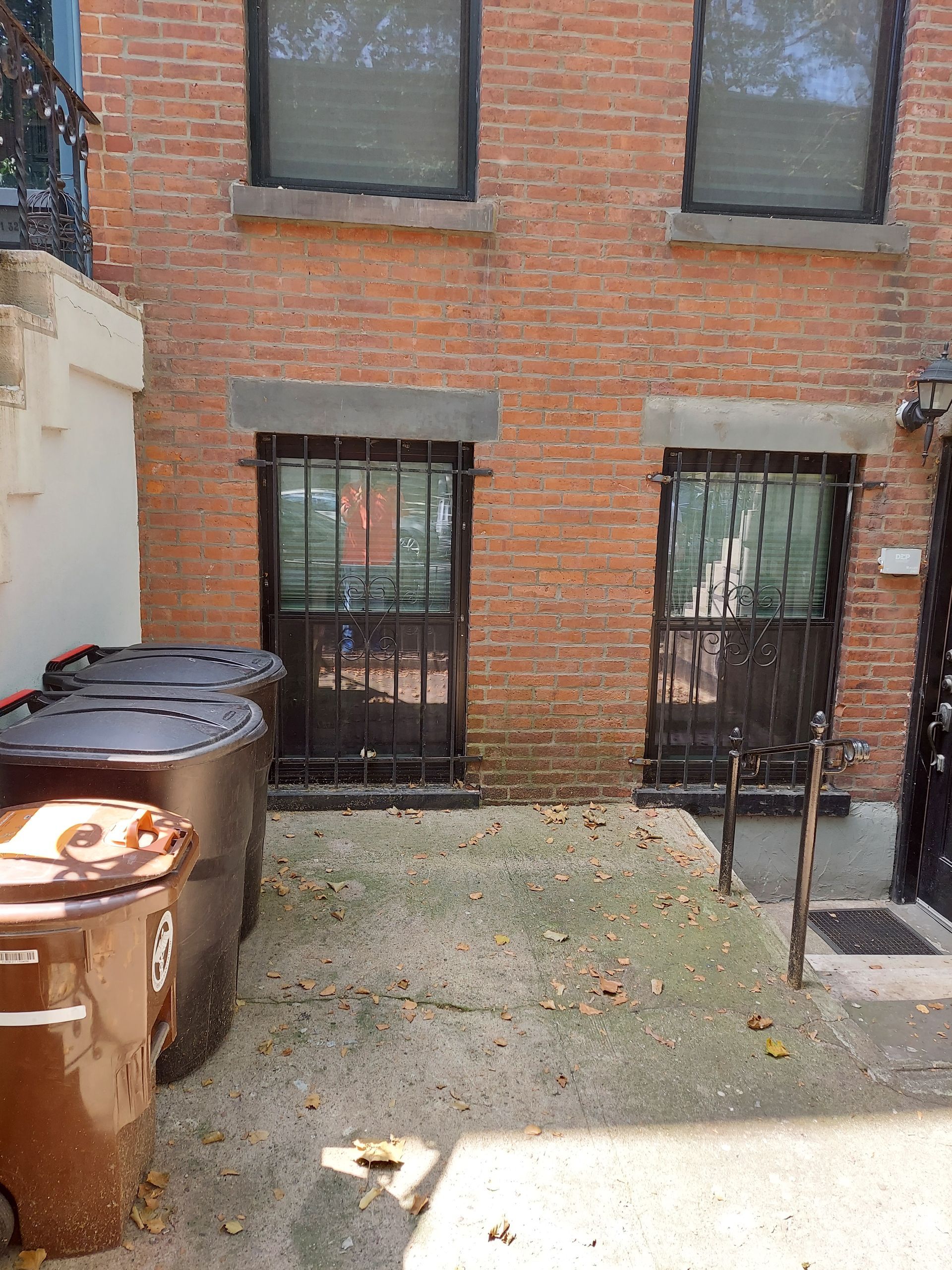 Brick courtyard with two barred basement doors, trash bins on the left, and a small paved area.