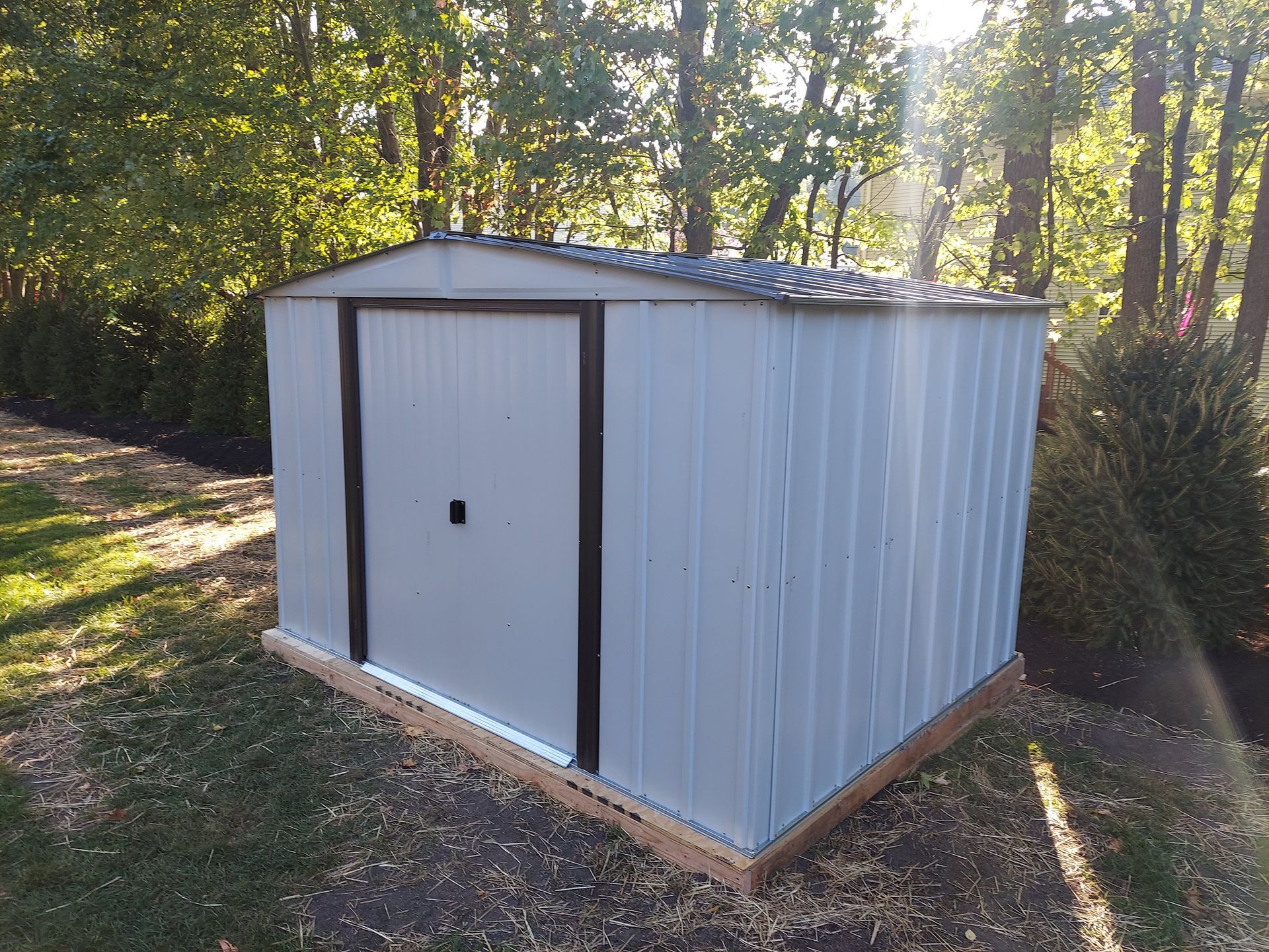 White metal storage shed with a single door in a wooded backyard.