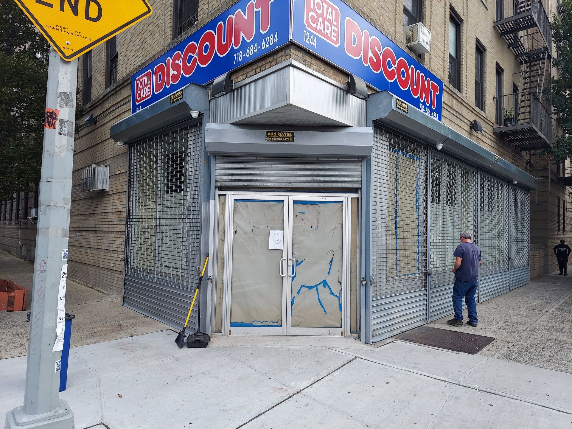 Closed discount store corner with metal shutters, glass doors, and a man standing on the sidewalk.