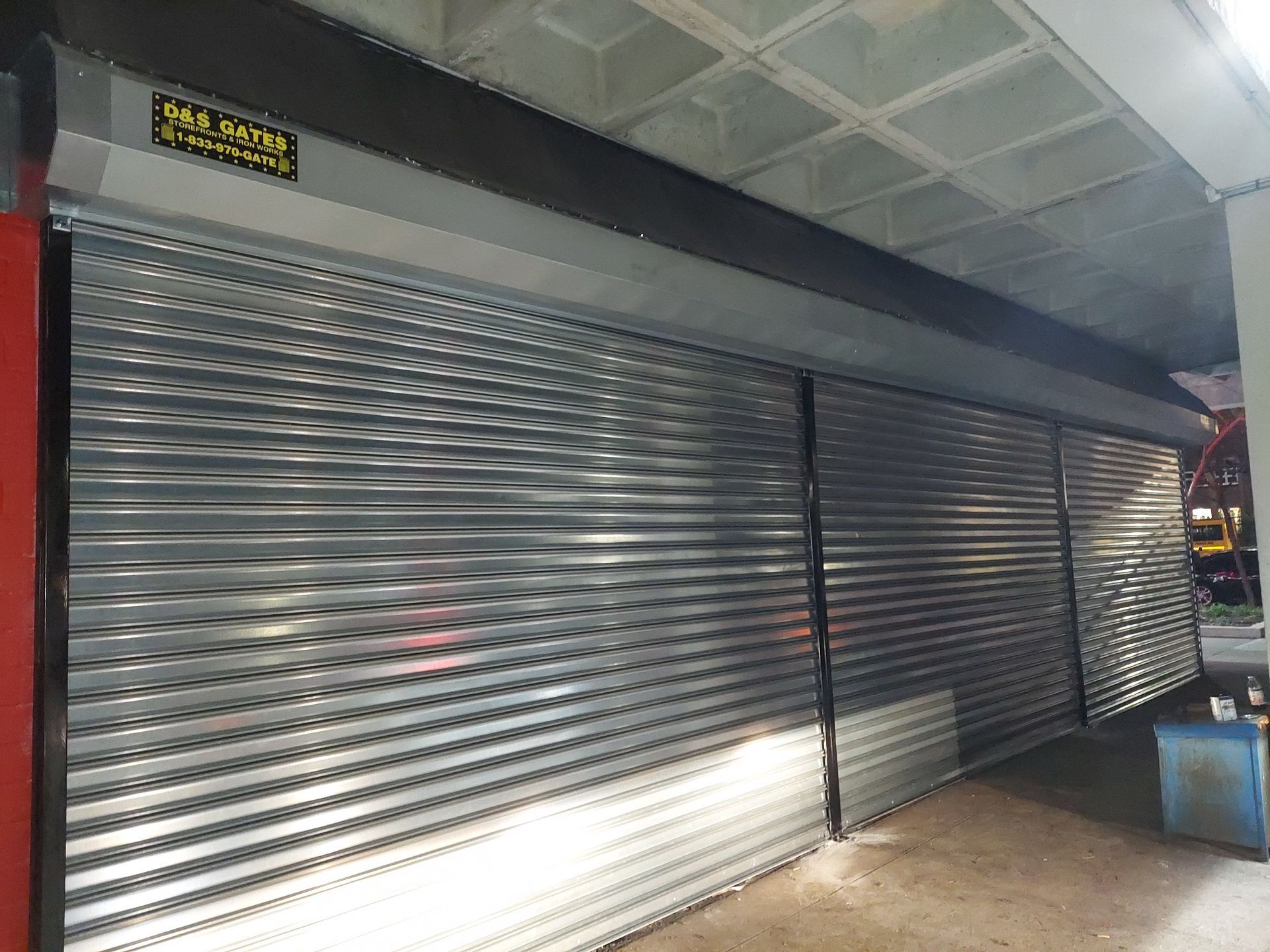 Closed metal roll-up shutters across a storefront in a covered walkway, with bright sunlight on the floor.